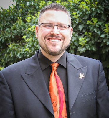 This is an image of a male person named Brown. He is wearing a black shirt with an orangish-red color tie and a Black suit. He is wearing eyeglasses. The photograph is taken in an outdoor environment with lots of leaves in the background.