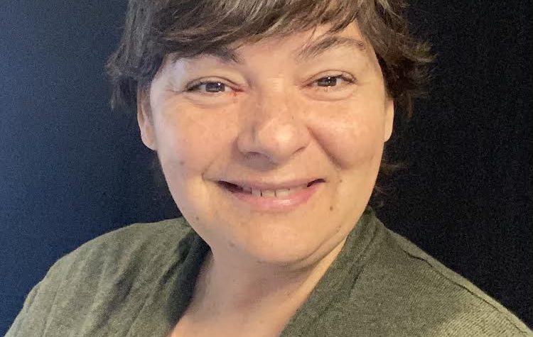 This is an image of a female person named Kim. she has short light brown hair. This is a closeup photograph of her and she is wearing a grey top with a pastel green jacket on top. The background is black.