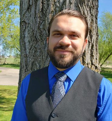 This is an image of a Male person named Marcus. He had a beard with well-combed hair. He is wearing a Blue shirt with a tie and a waistcoat. This image is clicked outdoors with a large tree behind him.