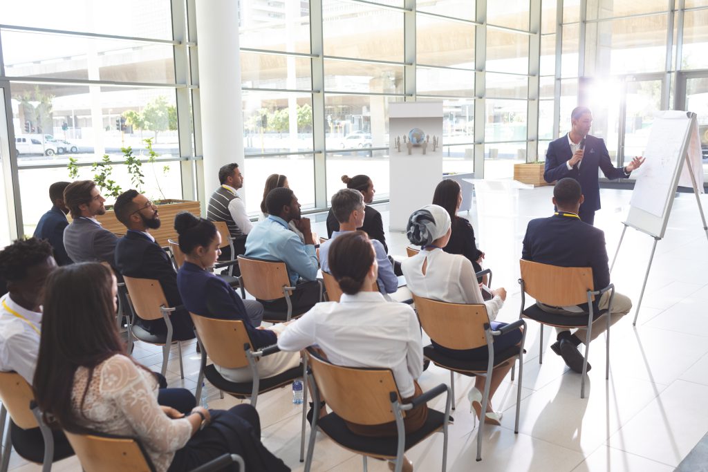 This is an image of a presentation. The presenter is having a mic in his hand and is the point to a board and appeared to be talking. There is an audience sitting in chairs. This is happening in a room with glass walls with bright light coming inside.
