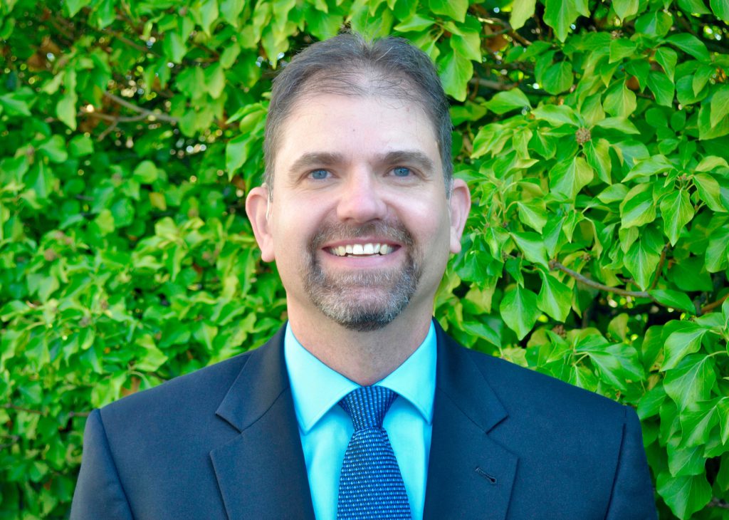 This is an image of a male person named " Dr. Wilson". He is wearing a light blue shirt, a blue dotted tie, and a blue suit. He has a french beard and mustache. The photograph is in an outdoor environment with leaves in the background.