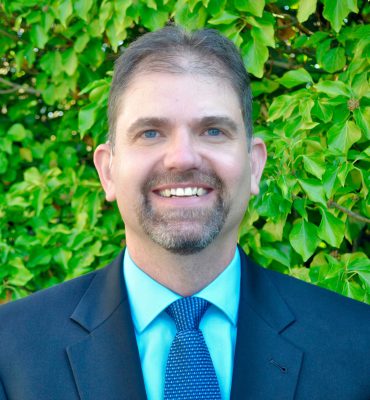 This is an image of a male person named " Dr. Wilson". He is wearing a light blue shirt, a blue dotted tie, and a blue suit. He has a french beard and mustache. The photograph is in an outdoor environment with leaves in the background.