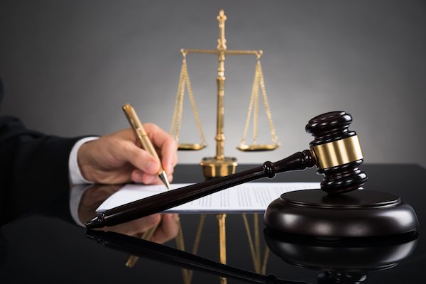 This image shows a black shiny table with a gavel, golden color scales of justice and a hand of a person in a suit with a golden pen in the hand that appears to be signing on some document kept on the table.