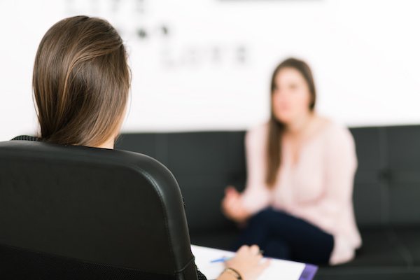 This image appears to be from a therapy session. In the background, there is a blurred image of a woman sitting on the couch facing another woman who appears to have a notepad and a pen in her hands while sitting in a black chair.