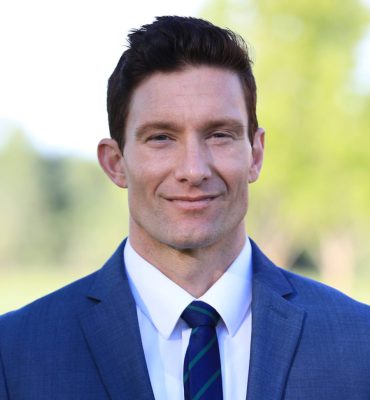 This image is of a male person named "Brandon Henscheid". He is cleaned and shaved with maintained black hair. He is wearing a White shirt, a Blue tie, and a Blue Suit. The background is outdoors.