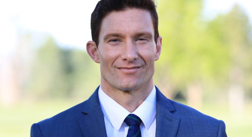This image is of a male person named "Brandon Henscheid". He is cleaned and shaved with maintained black hair. He is wearing a White shirt, a Blue tie, and a Blue Suit. The background is outdoors.