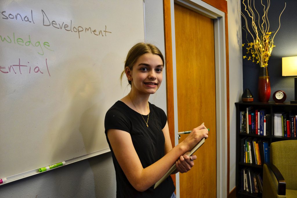 This image shows a young teenage girl wearing a blacktop and neck chain standing in front of a whiteboard with a pen and writing pad in her hands. on the board behind her, there is some text like "personal development", Knowledge". On the right side, there is a book stand with books inside, with a table lamp, vase, table clock, and showpiece kept on top of the book stand. Next to the bookstand, there is a brown door.