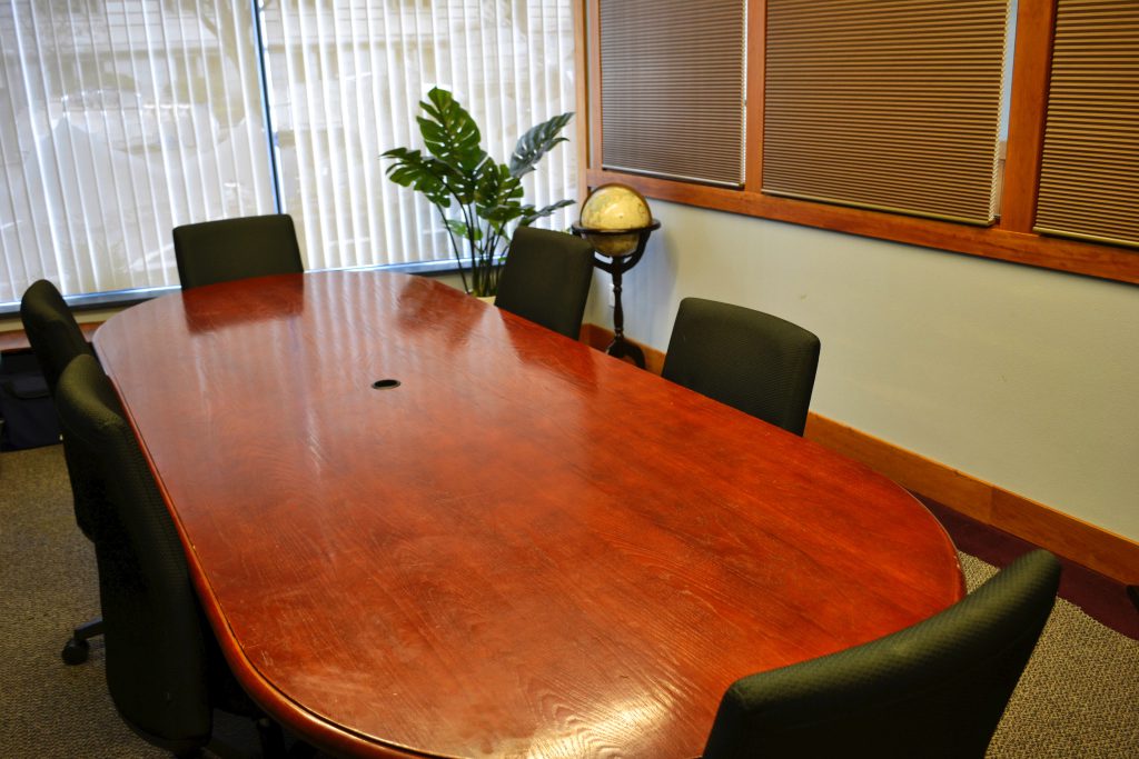This image appears to be of a conference room. There is are large rectangular conference table with round edges. There is a total of six chairs around the conference table. There is a glass window with blinds on it. There is a globe kept on the wooden stand next to one of the chairs.