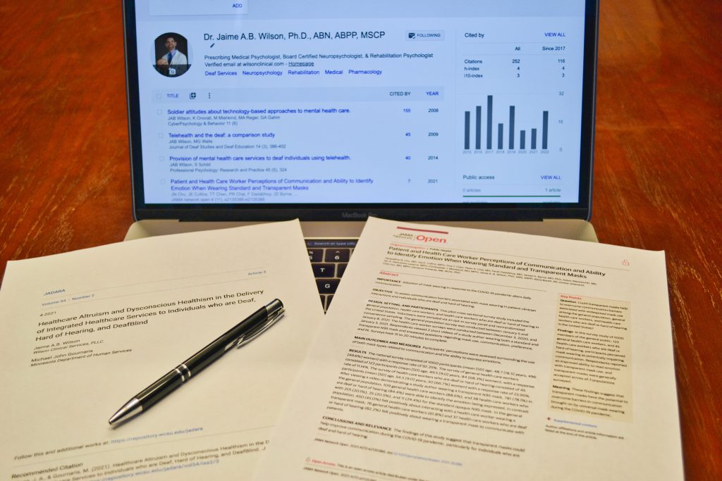 This image is of a screen of a laptop with some details about Dr. Jaime A B. Wilson, Ph.D., ABN, ABPP, MSCP. There are some bar graphs & some stats. on the laptop keyboard, there are paper printouts and a black pen on top of them. The paper has some articles about Healthcare Altruism and Dysconsious Healthism in the delivery of integrated healthcare services to individuals who are deaf, hard of hearing, and deafblind".